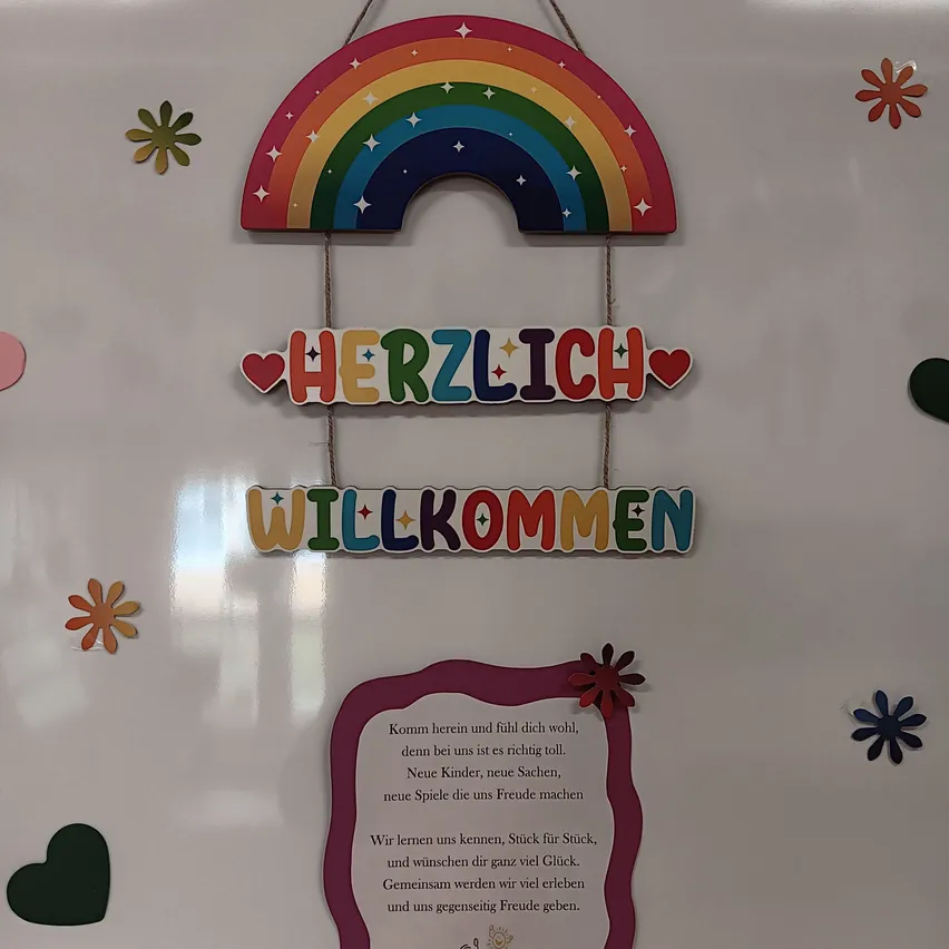 Ein farbenfrohes Regenbogenschild mit den Worten 'Herzlich Willkommen' hängt an einer weißen Wand, begleitet von dekorativen Blumen und Herzen. Darunter befindet sich eine Nachricht auf Deutsch, die neue Kinder, Gegenstände und Spiele einlädt, sich anzuschließen und Freude zu bringen.