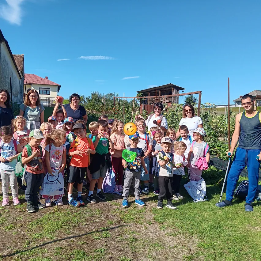 Eine Gruppe von Kindern und Erwachsenen steht in einem Garten, einige halten Ballons und Äpfel, unter einem klaren blauen Himmel.