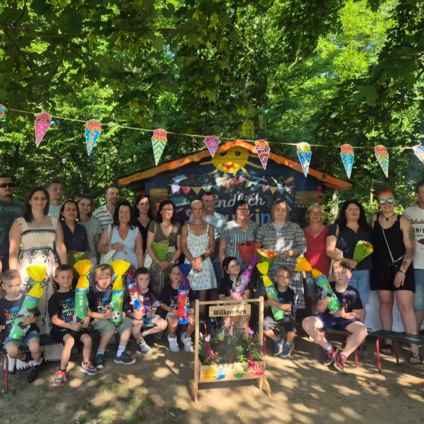 Eine Gruppe von Erwachsenen und Kindern posiert für ein Foto in einem Park, einige halten Blumen und Ballons, unter dekorativen Bannern und einer sonnenthematischen Struktur.