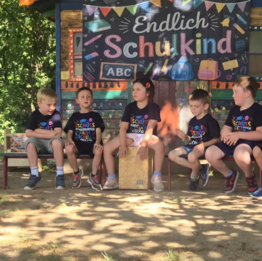 Fünf junge Kinder sitzen vor einer farbenfrohen Kulisse, die 'Endlich Schulkind' mit verschiedenen aufgezeichneten Schulartikeln zeigt.