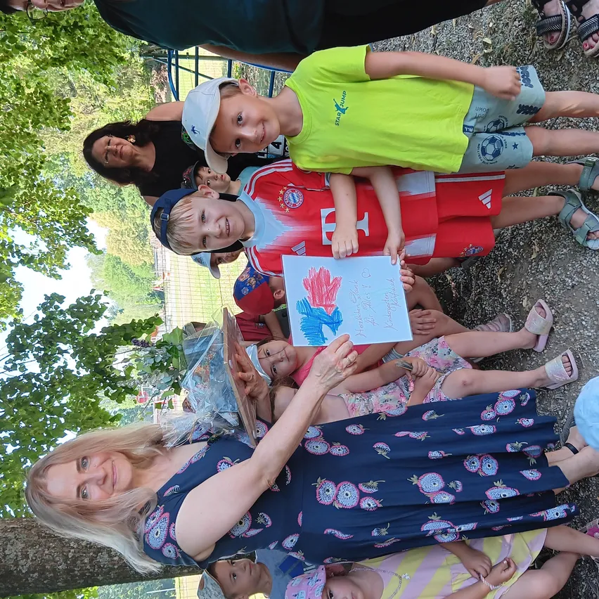 Eine Gruppe von Kindern und Erwachsenen sitzt auf dem Boden in einem Park. Eine Frau hält ein Papier mit blauen Handabdrücken. Hinter ihr stehen mehrere Kinder, eines hält einen Blumenstrauß. Sie sind von Grün umgeben.