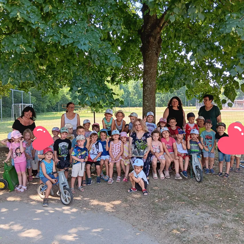 Eine Gruppe von Kindern und Erwachsenen posiert unter einem Baum in einem Park. Einige halten Blumen, und zwei große Herzen sind im Vordergrund.