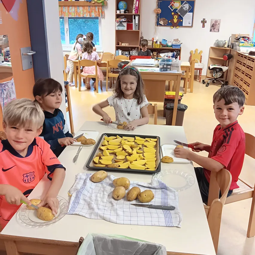 Vier Kinder sitzen um einen Tisch in einem Klassenzimmer, jeder mit einem Teller geschnittener Kartoffeln. Ein Mädchen in der Mitte hält ein Messer und schneidet Kartoffeln.