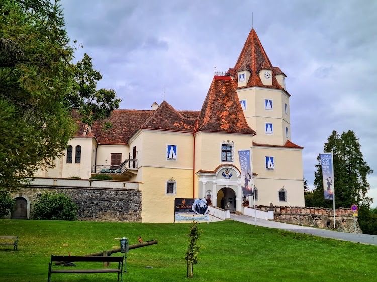 Ein großes historisches Gebäude mit rotem Dach und Uhrturm steht in einem Park mit grünem Rasen und Bäumen.