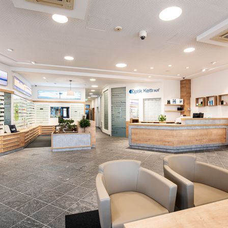 Innenraum eines Optikers mit modernen Möbeln, Holztheke und dekorativen Pflanzen. Die Decke hat runde Lichter, und Regale zeigen verschiedene Gegenstände.