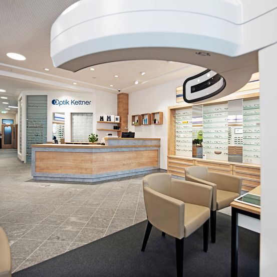 Der Innenraum eines Optikers zeigt einen hölzernen Empfangstresen, moderne Beleuchtung und Vitrinen mit Brillen. An der Wand steht der Name 'Optik Kettner' und ein Schild. Es gibt Stühle und einen schwarzen Teppich im Wartebereich.
