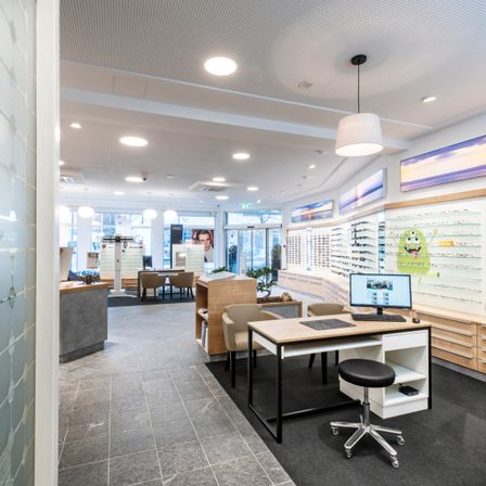 Das Innere eines Optikgeschäfts mit modernen Designelementen, einschließlich eines Schreibtisches mit Monitor, einem Hocker und mehreren Stühlen. Es gibt Glasvitrinen mit Brillen und gerahmte Poster an der Wand.