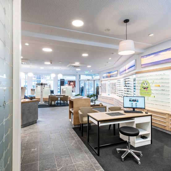 Das Innere eines Optikgeschäfts mit modernen Designelementen, einschließlich eines Schreibtisches mit Monitor, einem Hocker und mehreren Stühlen. Es gibt Glasvitrinen mit Brillen und gerahmte Poster an der Wand.
