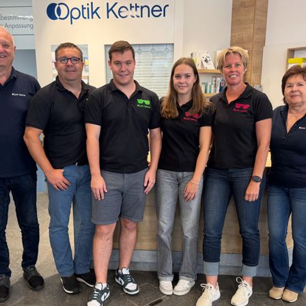 Eine Gruppe von sechs Personen, die schwarze Hemden mit dem Wort Optik Kettner tragen, steht in einem Geschäft zusammen. Sie lächeln und posieren für ein Foto.