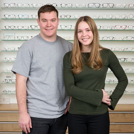 Ein Mann und eine Frau stehen lächelnd vor einer Wand mit vielen ausgestellten Brillenpaaren.