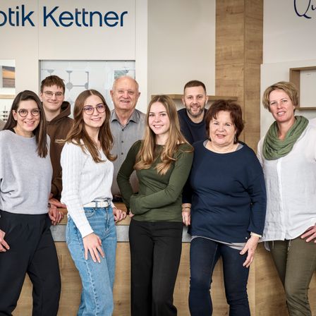 Eine Gruppe von Menschen steht vor einem Schild mit der Aufschrift Optik Kettner. Sie lächeln und scheinen für ein Foto zu posieren.