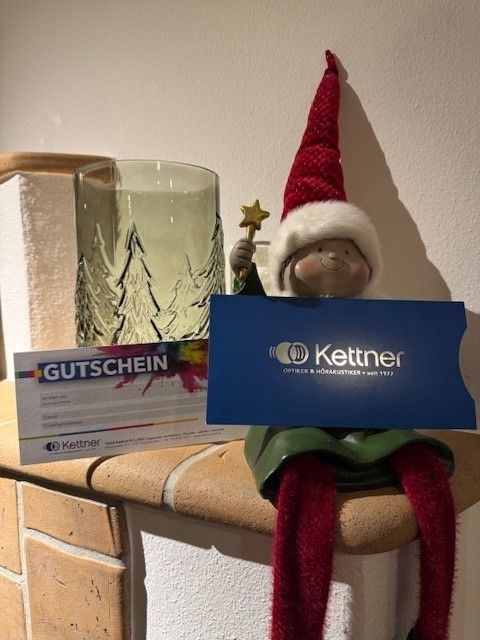 Ein kleiner Gnom hält eine blaue Kettner-Geschenkkarte, mit einem Glasgefäß dahinter und einer weißen Schachtel links.