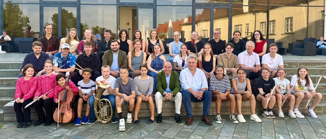 Eine Gruppenaufnahme von Musikern und Kindern vor einem Gebäude. Einige sitzen, andere stehen. Instrumente wie Cello und Tuba sind sichtbar.
