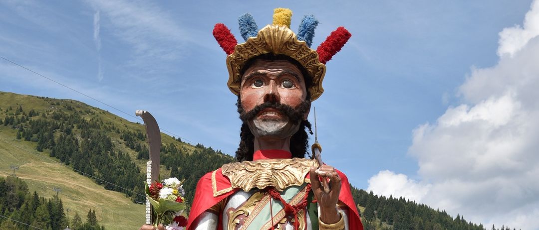 Ein großer Mann, gekleidet in einen goldenen und roten Kilt und mit einem Blumenstrauß in der Hand, steht vor einem Berg.