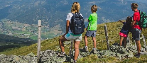 Eine vierköpfige Familie steht auf einem felsigen Hügel und blickt auf ein Tal und eine Bergkette mit bewölktem Himmel. Sie tragen alle Rucksäcke.