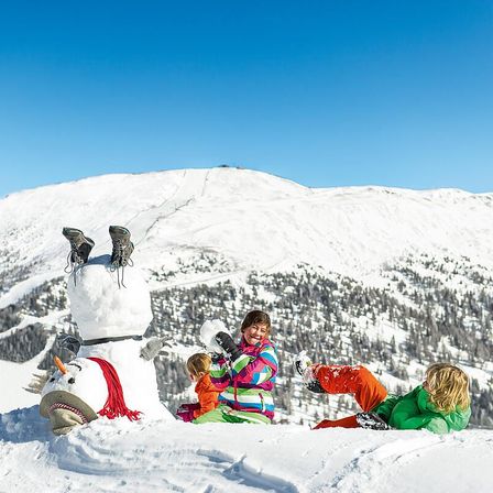 Eine dreiköpfige Familie in Winterkleidung posiert neben einem Schneemann auf einem verschneiten Berg, wobei ein Kind eine Schneeball hält.