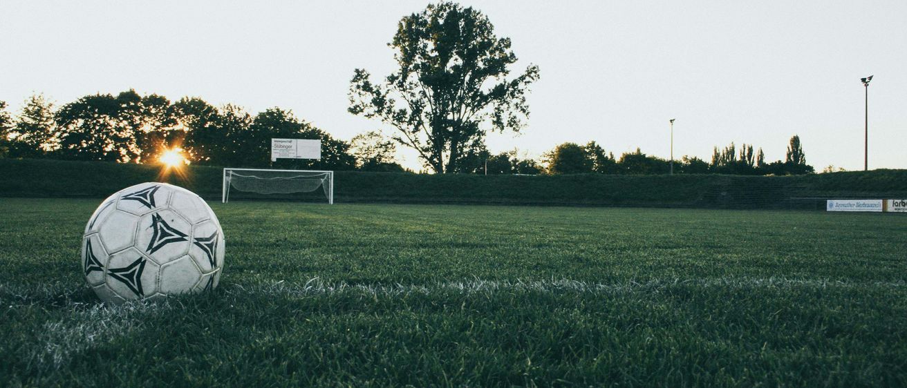 Ein Fußball liegt auf einem Feld mit einem Tor am Horizont, umgeben von Bäumen und einem klaren Himmel.