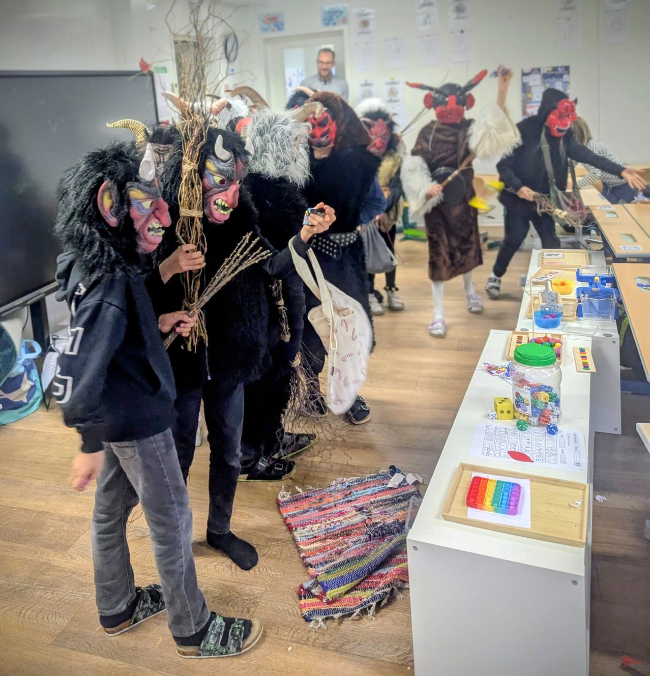 Mehrere Menschen in Hörner- und Maskenkostümen stehen in einem Klassenzimmer und halten Requisiten. Ein Tisch mit Würfeln, einem Glas und einem Teppich ist sichtbar. Eine weitere Person steht in der Nähe einer Tafel.