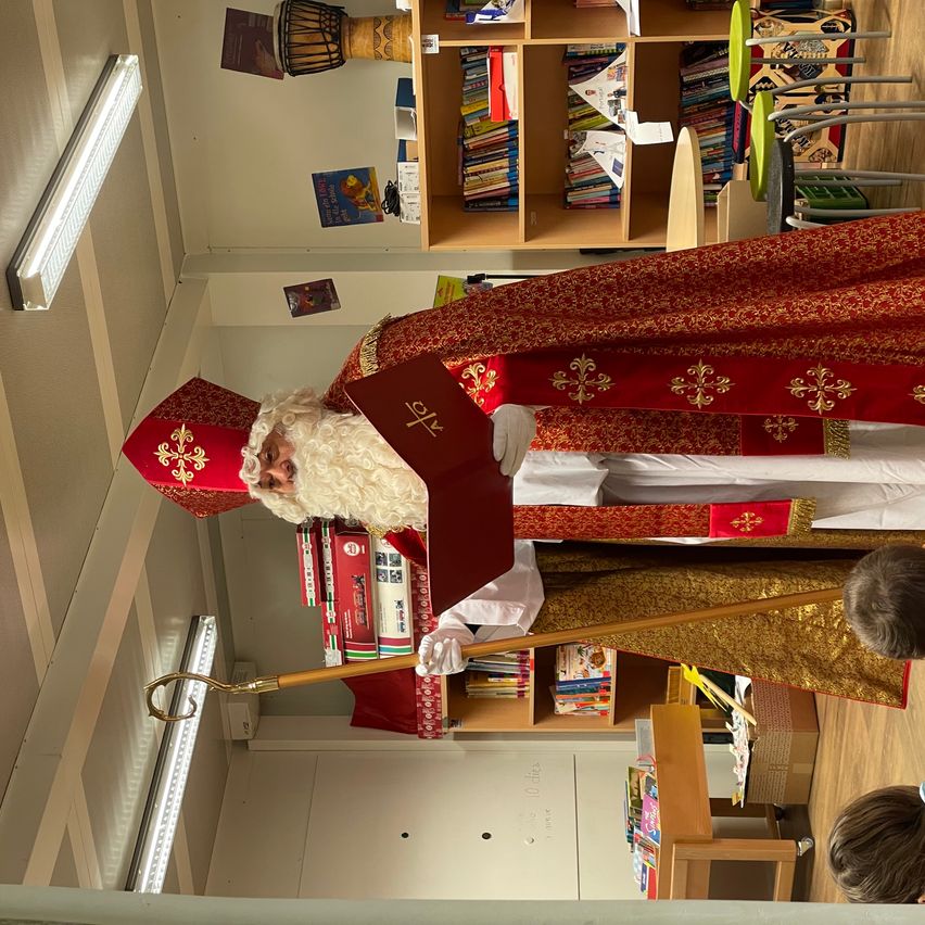 Eine als Weihnachtsmann verkleidete Person mit Buch und Stab steht in einem Raum mit Bücherregalen und einem beobachtenden Kind.