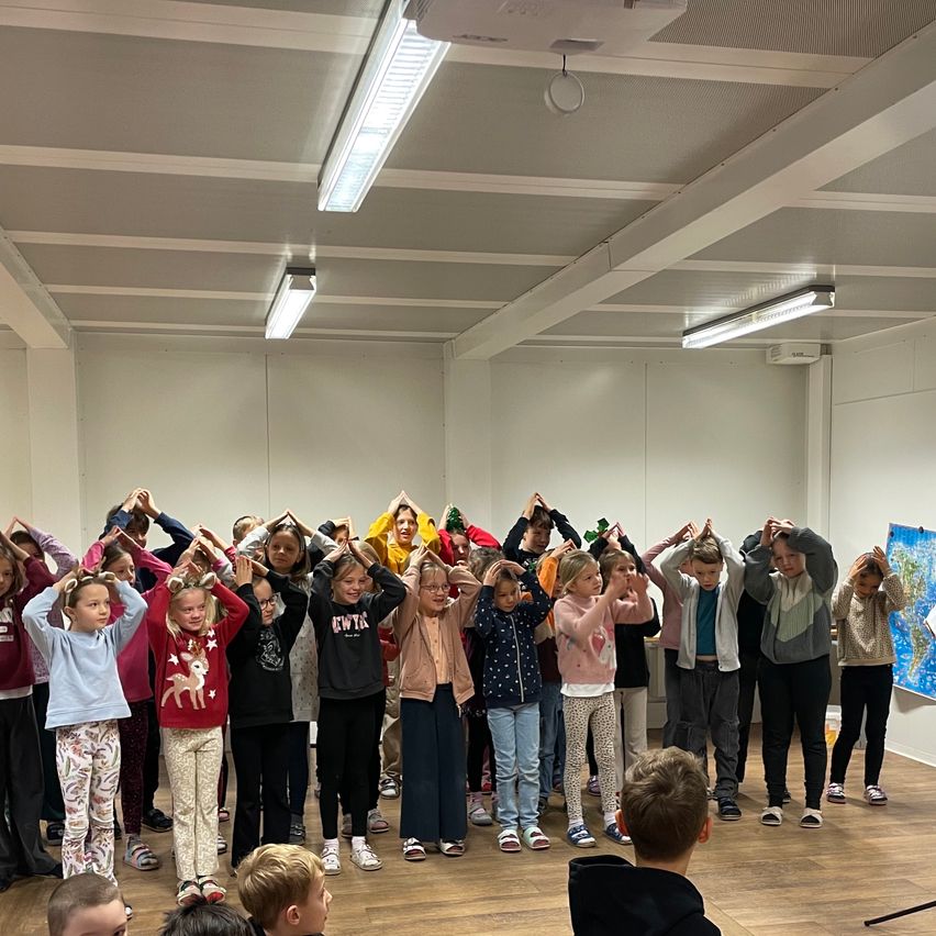 Eine Gruppe von Kindern und Jugendlichen steht im Kreis mit den Händen auf dem Kopf, wahrscheinlich üben sie eine Tanz- oder Übungsroutine in einem Raum mit Holzboden und weißen Wänden.