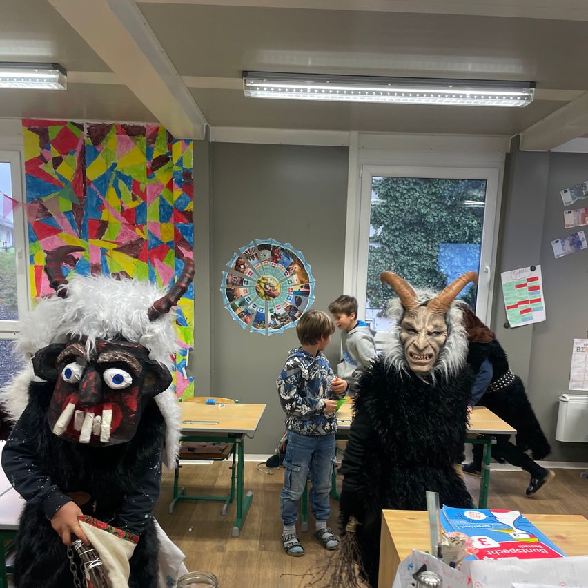 Ein Klassenzimmer mit Kindern in Kostümen, einer hält eine Tasche. Ein rundes Kunstwerk an der Wand, Tische und eine Tür mit einem Glasfenster.