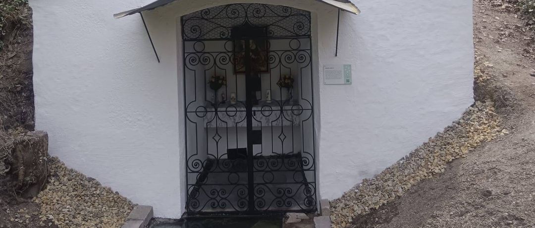 Eine kleine weiße Kapelle mit einem Glockenturm und einem Kreuz oben. Sie hat einen gewölbten Eingang mit einem Metalltor. Im Inneren befindet sich ein weißer Tisch mit Blumen und einem Schild.