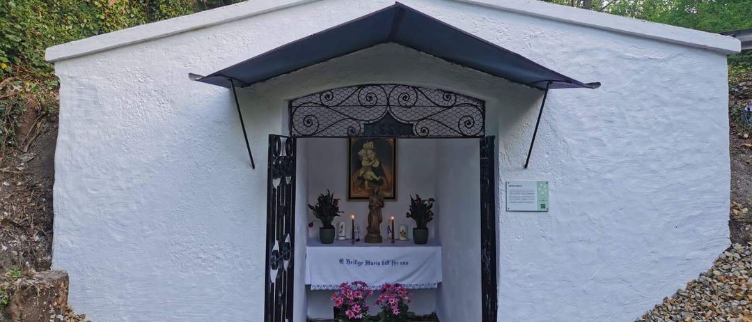 Eine kleine weiße Kapelle mit einem Kreuz auf der Spitze und einem schwarzen Vordach. Im Inneren befindet sich ein Altar mit einer Kerze, Blumen und einem Bilderrahmen.