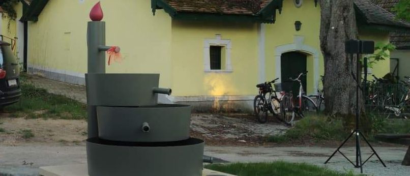 Ein Priester in Weiß steht neben einem Brunnen, mit einem gelben Haus, Bäumen und einem schwarzen Auto im Hintergrund. In der Nähe sind Fahrräder und ein Stativ.