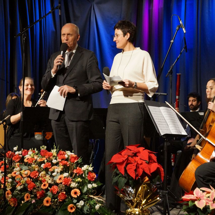 Ein Mann und eine Frau stehen auf der Bühne und halten Mikrofone. Hinter ihnen spielt eine Frau Geige und ein Mann Cello. Musikinstrumente und Notenblätter sind auf der Bühne.