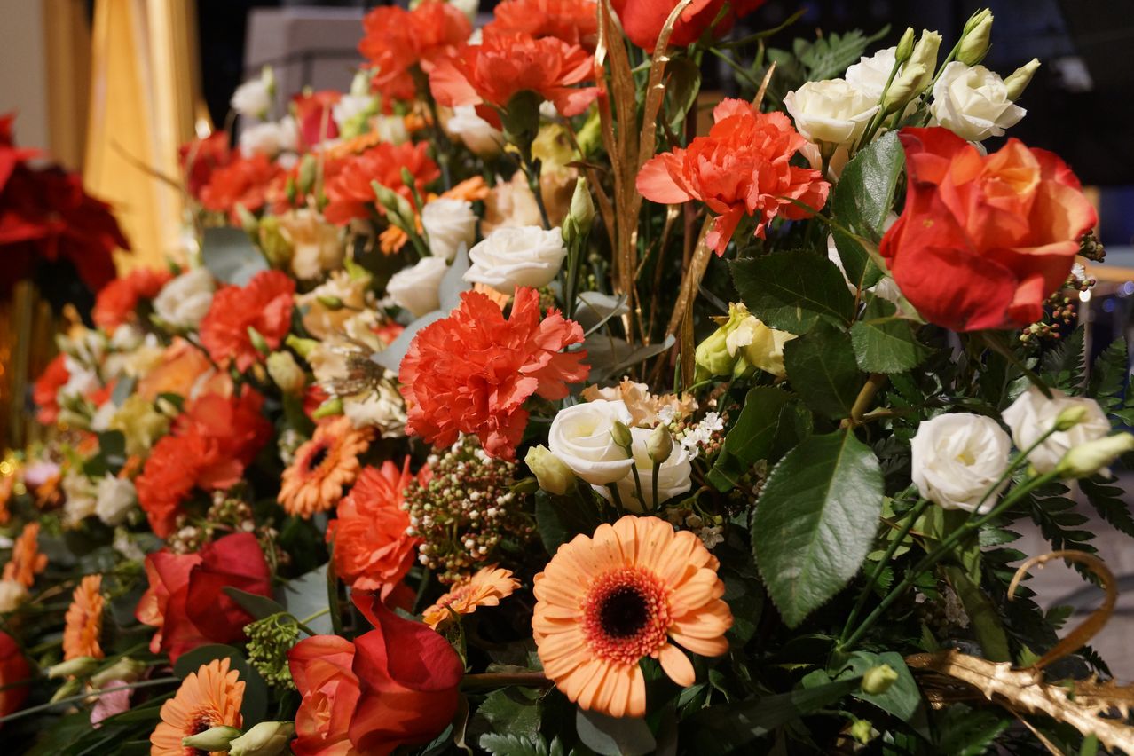 Eine farbenfrohe Blumenarrangement enthält rote, orange und weiße Blumen, wobei einige Blätter und Stängel sichtbar sind. Im Mittelpunkt steht eine große orange Daisy mit einem prominenten roten Zentrum.