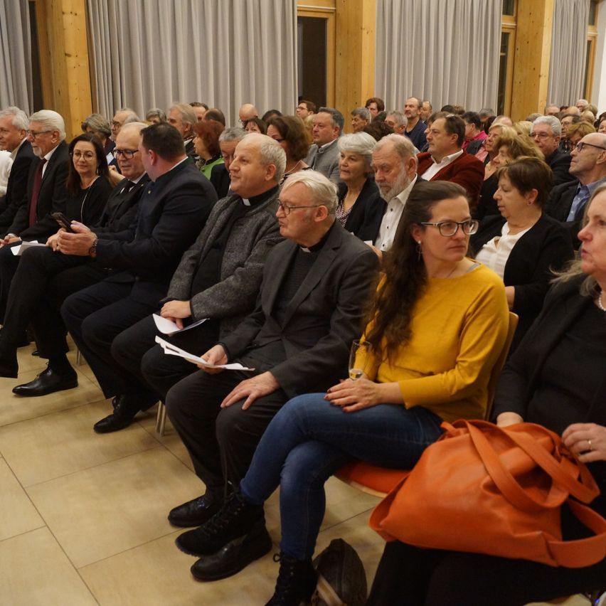 Eine große Versammlung von Menschen sitzt in Reihen, vermutlich in einem Konferenzsaal. Einige lesen Papiere, während andere in Gespräche vertieft sind. Eine Frau in einem gelben Pullover hält ein Weinglas. Eine Person in der vorderen Reihe benutzt ein Handy.