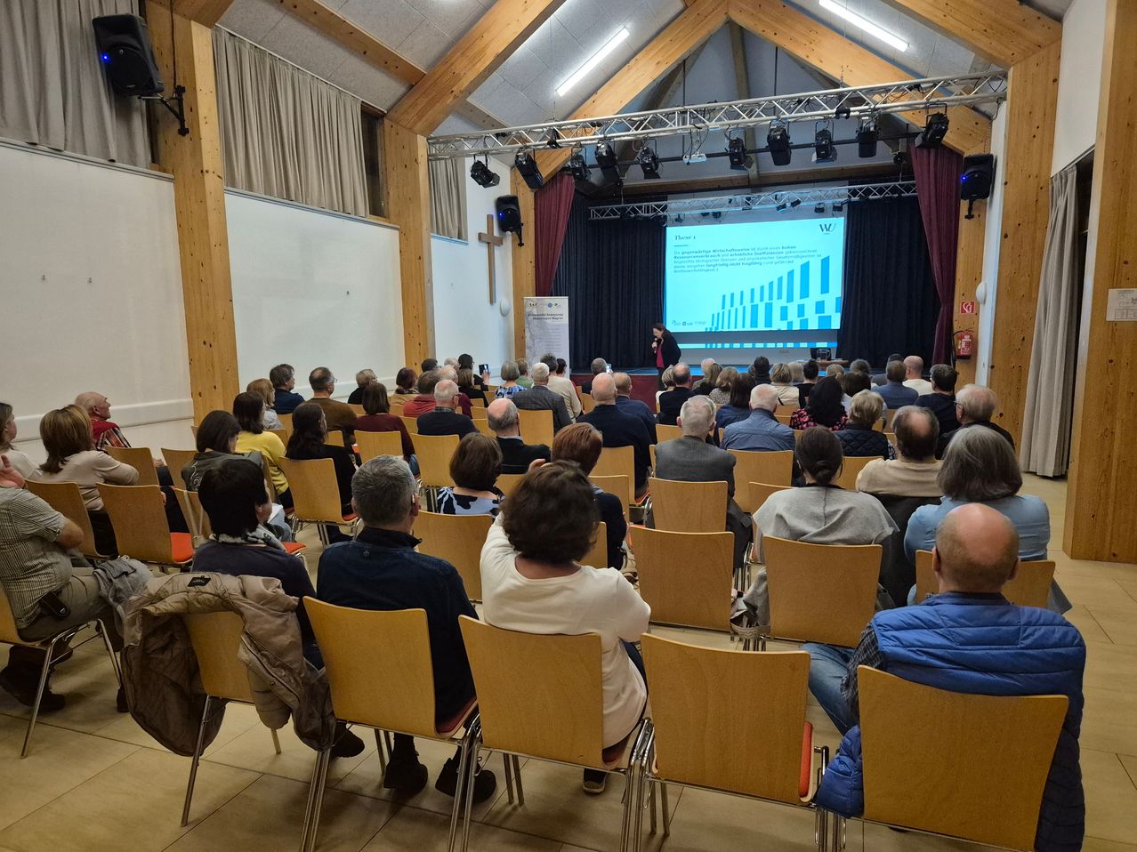 In einem Saal mit Holzwänden findet eine Vorlesung statt. Eine Person steht auf der Bühne und hält eine Präsentation mit einem Projektionsbildschirm. Das Publikum sitzt auf Stühlen, einige von ihnen tragen Masken.