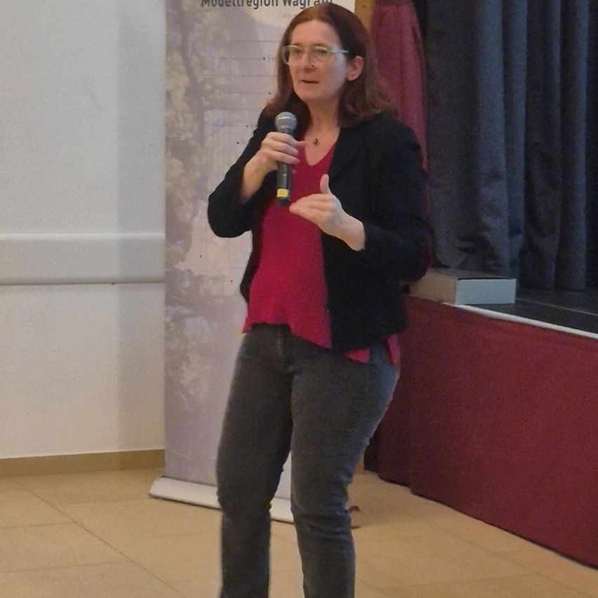 Eine Frau mit Brille spricht in ein Mikrofon und gestikuliert mit den Händen. Sie trägt ein rotes Hemd und eine schwarze Jacke und steht vor einem Banner.