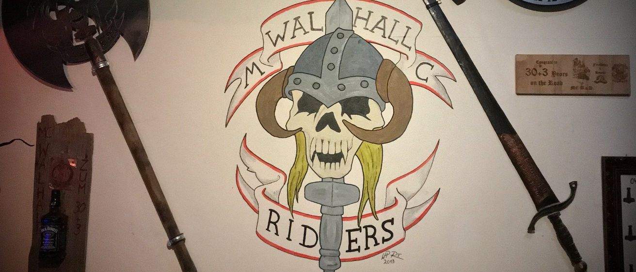 Eine Wand mit einem Schädel und Hörnern, der einen Helm trägt. Darunter steht der Text 'M Walhall Riders.' Es gibt zwei Schwerter und eine Axt an der Wand.