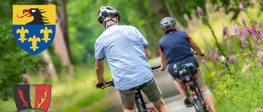 Zwei Radfahrer fahren auf einem Schotterweg durch einen üppigen grünen Wald. Sie tragen Helme und fahren Fahrräder. Wappen sind auf der linken Seite des Bildes angebracht.