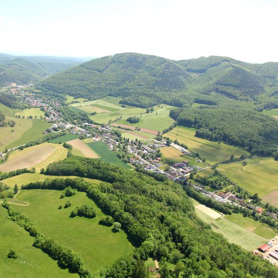 Luftaufnahme eines Dorfes inmitten einer üppigen grünen Landschaft, umgeben von Ackerland und dichten Wäldern. Die Landschaft zeigt sanfte Hügel und eine ruhige, malerische Umgebung.