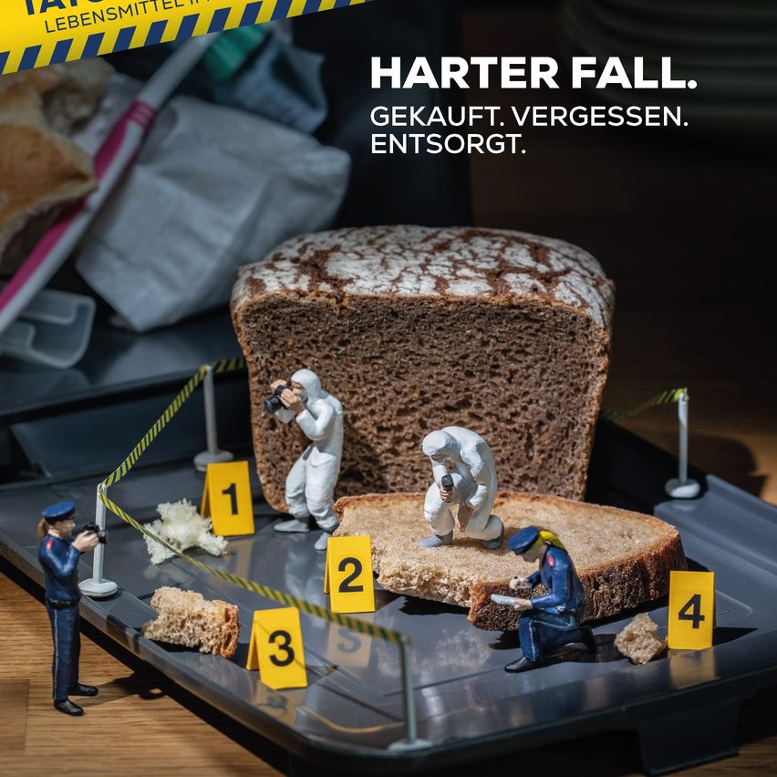 Eine Szene mit Miniatur-Polizeibeamten, die eine Kriminalszene untersuchen. Ein Laib Brot ist prominent in Szene gesetzt, mit umherliegenden Krümeln und nummerierten Beweismittelmarkierungen. Ein Beamter macht ein Foto, während andere das Brot untersuchen.