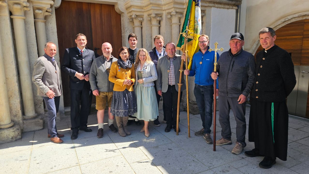 Eine Gruppe von Menschen steht vor einer Kirche und lächelt für ein Foto. Sie halten Flaggen und tragen verschiedene Kleidungsstücke.