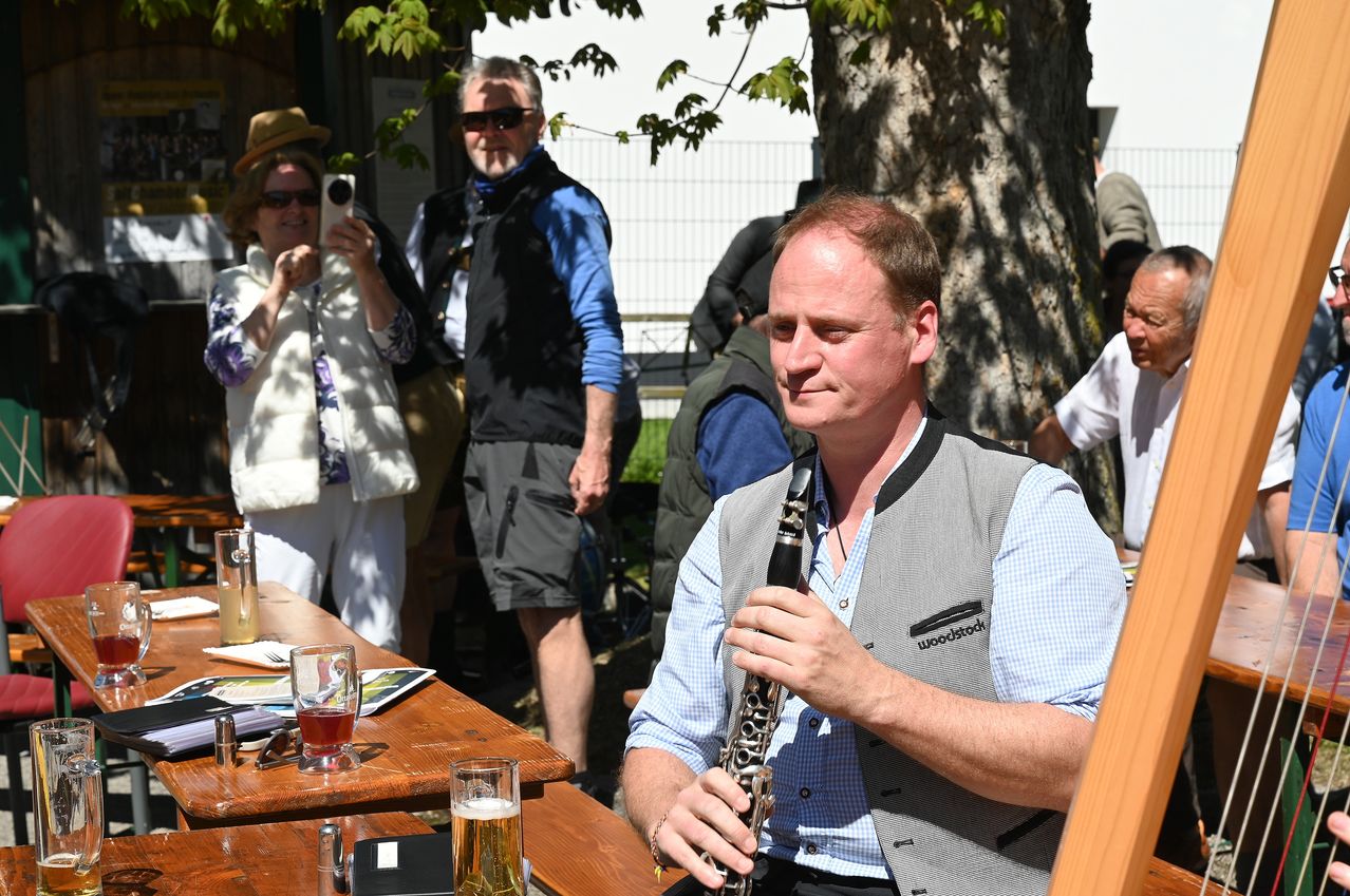 A man in a blue shirt plays a clarinet at an outdoor table, where glasses of beer are set. Behind him, a woman takes a picture. Another person stands near a tree.