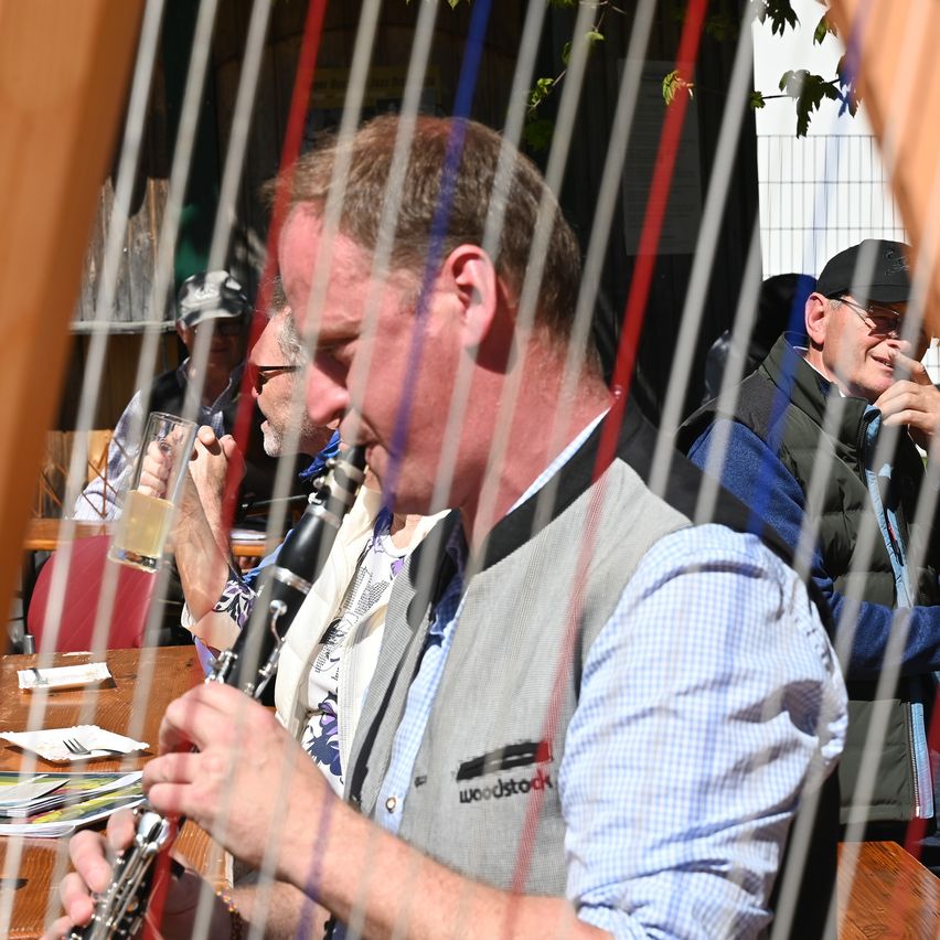 A man plays a clarinet at a restaurant. Other men sit behind him, one wearing glasses. Behind them, a wooden fence is visible.
