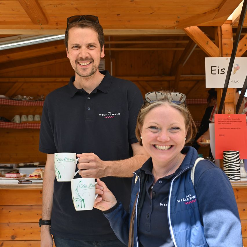 A man and woman smiling and posing for a photo while holding two mugs with the word 'Die' on them. Behind them is a wooden structure with a signboard with the word 'Eis'.