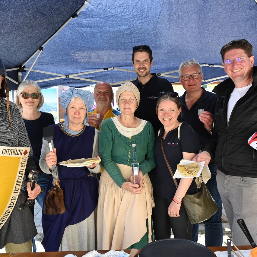 A group of people dressed in medieval clothing stand under a blue tent, posing for a photo. They hold food and drinks, with one person wearing a knight's helmet.
