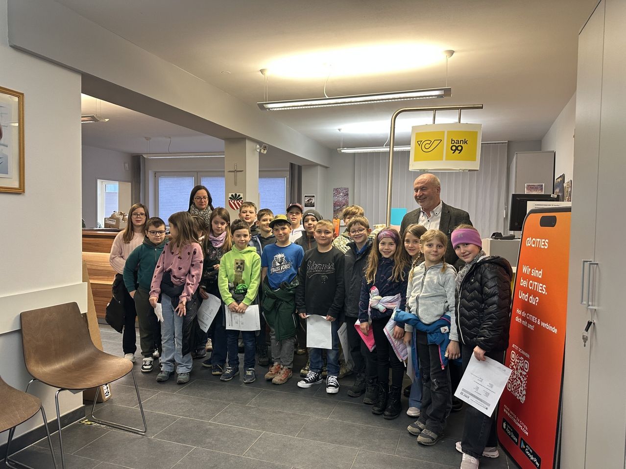 Eine Gruppe von Kindern und Erwachsenen steht in einer Bankhalle. Jedes Kind hält ein Stück Papier in der Hand. Ein erwachsener Mann lächelt und posiert mit den Kindern. Hinter ihnen steht ein Schild mit der Aufschrift 'Bank 99'.