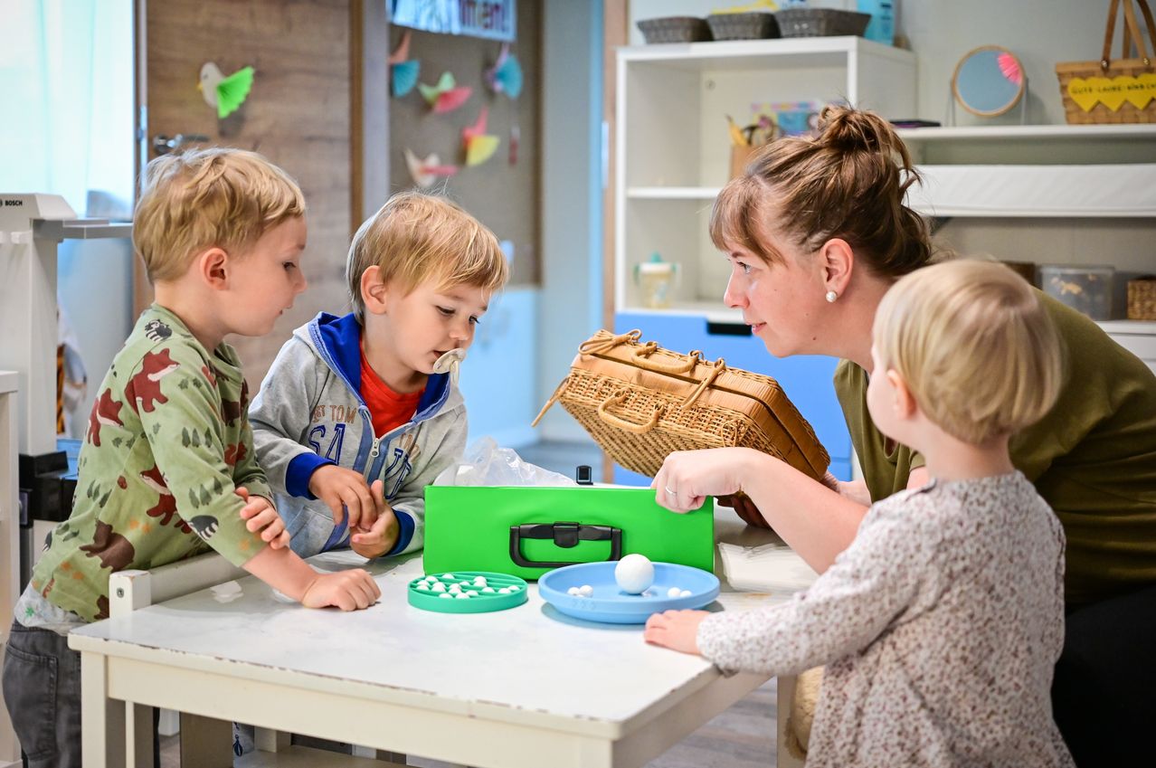 Eine Frau und drei Kinder sitzen an einem Tisch und spielen mit einer Tasche und einem Spielbrett. Die Frau schaut die Kinder an, während sie spielen.