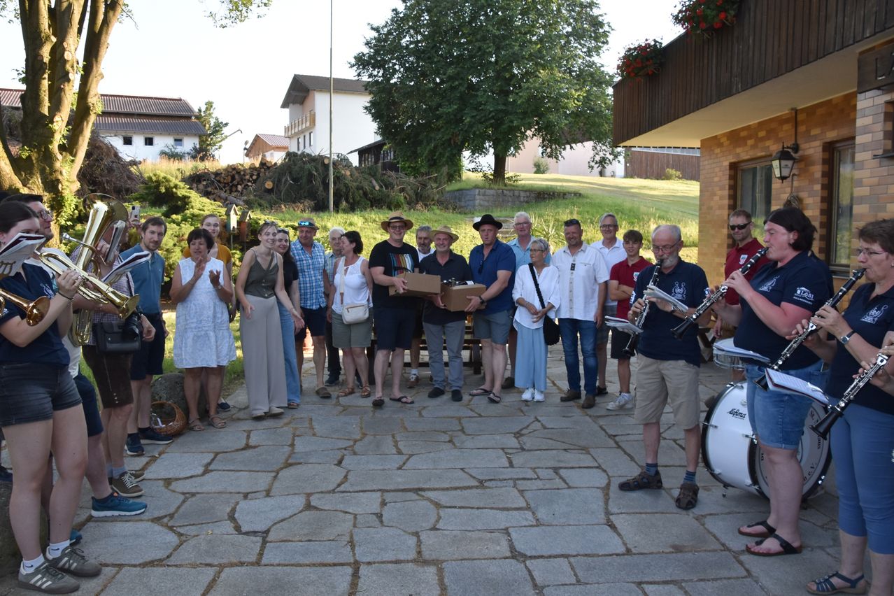 A group of people are gathered outside. One man holds a box, and a man on the right plays a clarinet. Another man on the right plays a drum. Some are wearing hats and glasses. Trees and a building are in the background.