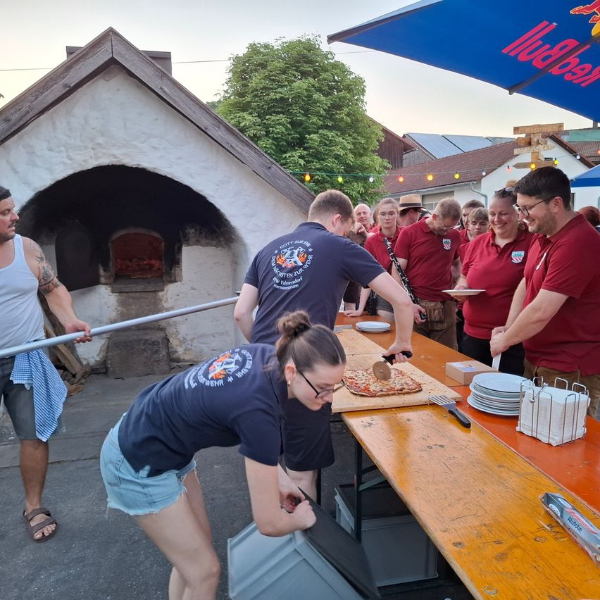 People in matching shirts prepare and serve pizza in a rustic setting with an outdoor oven. A table holds plates and pizza tools. A man holds a pizza peel and another slices pizza.