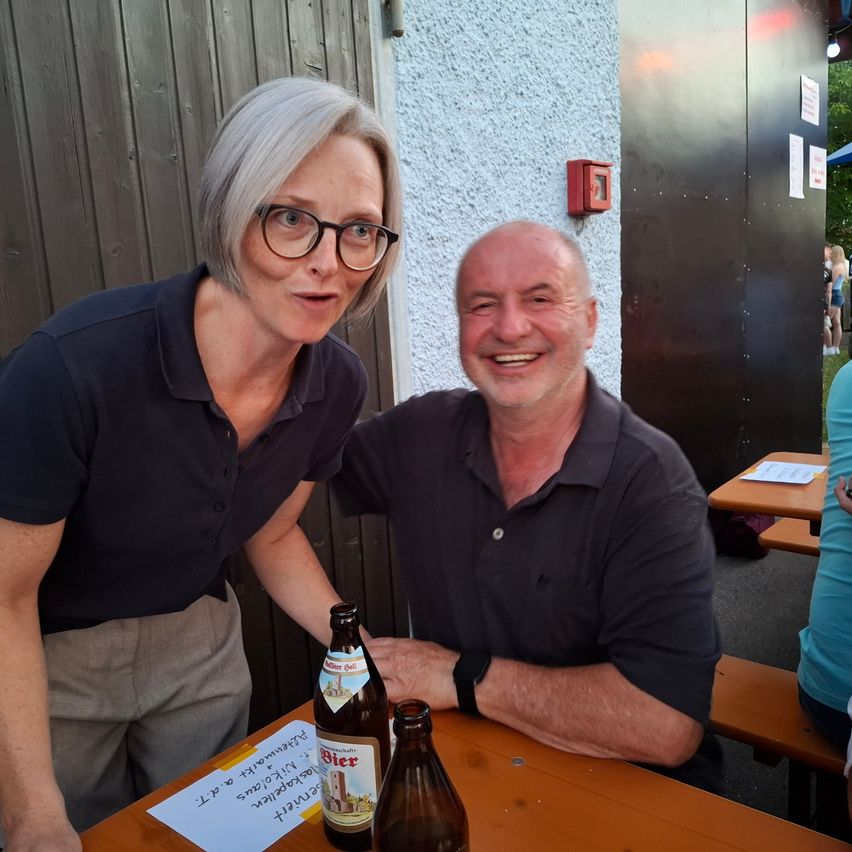 A man and woman sit at an outdoor table with bottles of beer in front of them. The woman wears glasses and a dark polo shirt, while the man has a bald head and wears a dark polo shirt and a black wristwatch. Behind them, there are tables and benches, with a wooden door and fire extinguisher visible.