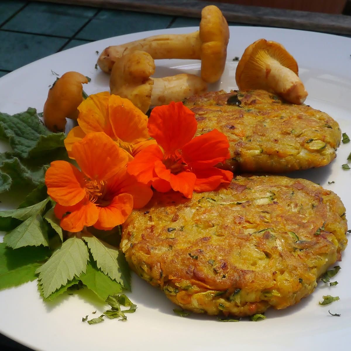 Ein weißer Teller mit zwei goldbraunen Gemüsepatties, verziert mit orangefarbenen Blumen und grünen Blättern, begleitet von einigen Pilzen.