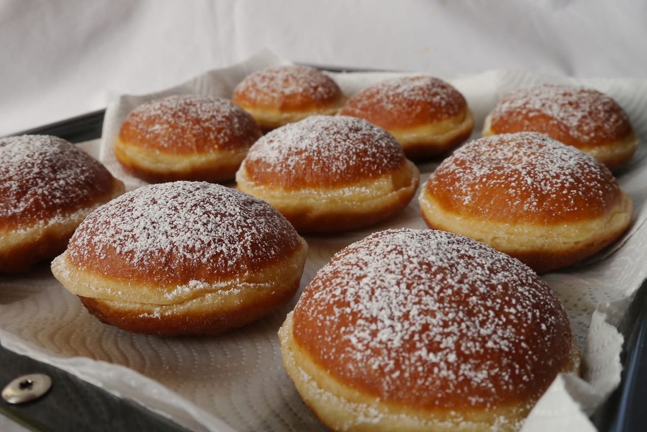 Ein Tablett mit frisch zubereiteten Donuts, die mit Puderzucker bestreut sind, auf einem weißen Tuch. Jeder Donut ist goldbraun und leicht aufgebläht.