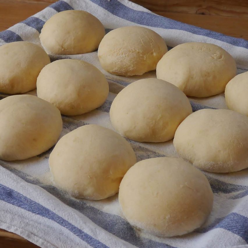Auf einem Holztisch liegt ein Handtuch, auf dem mehrere Teigkugeln gleichmäßig verteilt sind. Die Teigkugeln sind golden und sehen weich aus.
